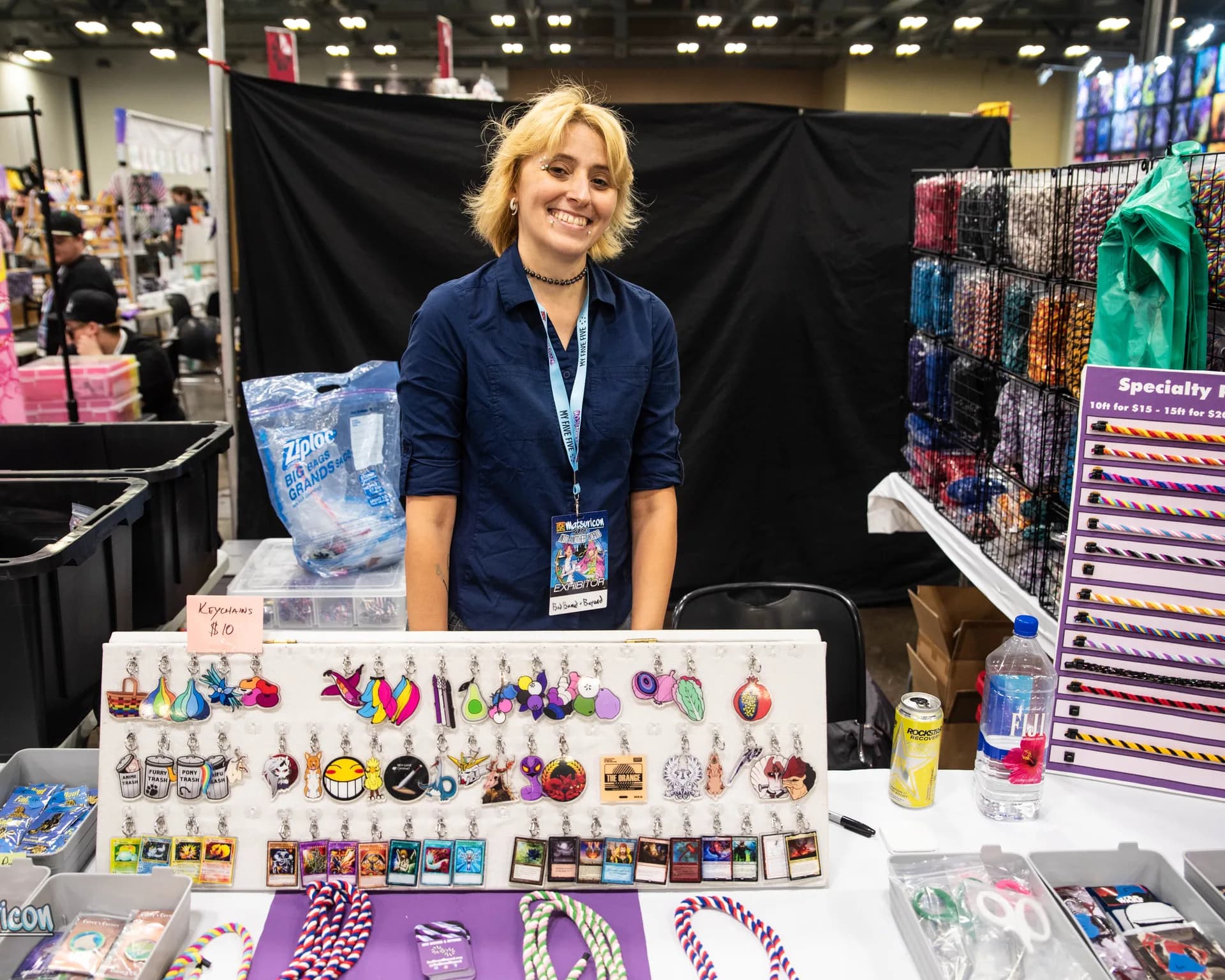 A smiling Matsuricon exhibitor stands behind a display of keychains, trading cards, and accessories.