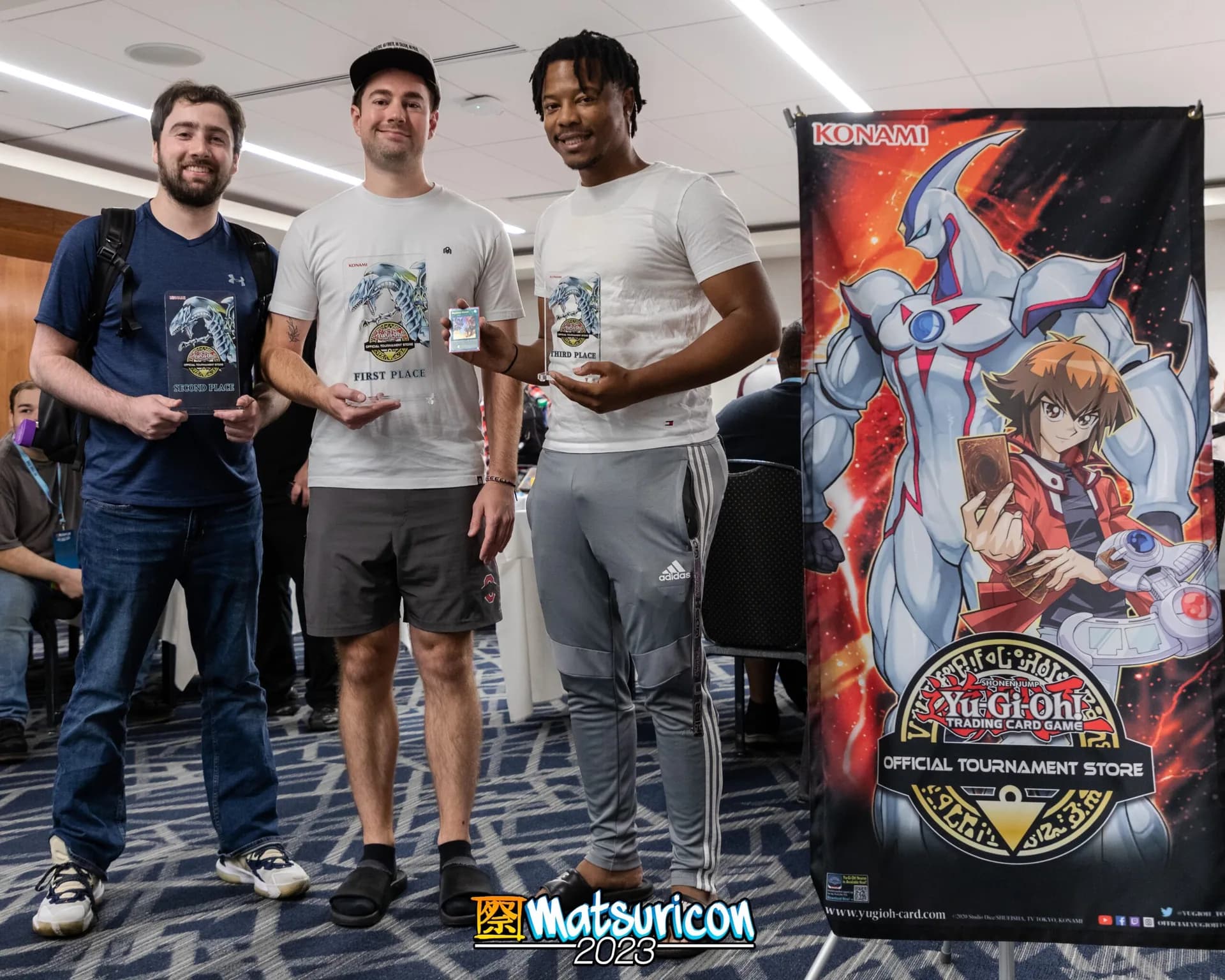 Three Yu-Gi-Oh tournament winners hold their prizes beside an official tournament banner.
