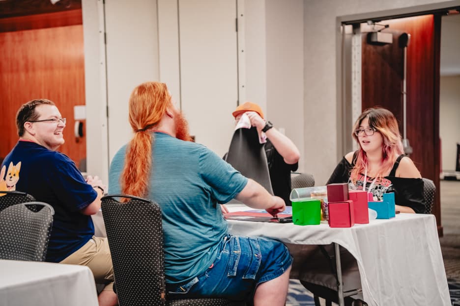 Four attendees smile and talk around a card game table in the CCG room.