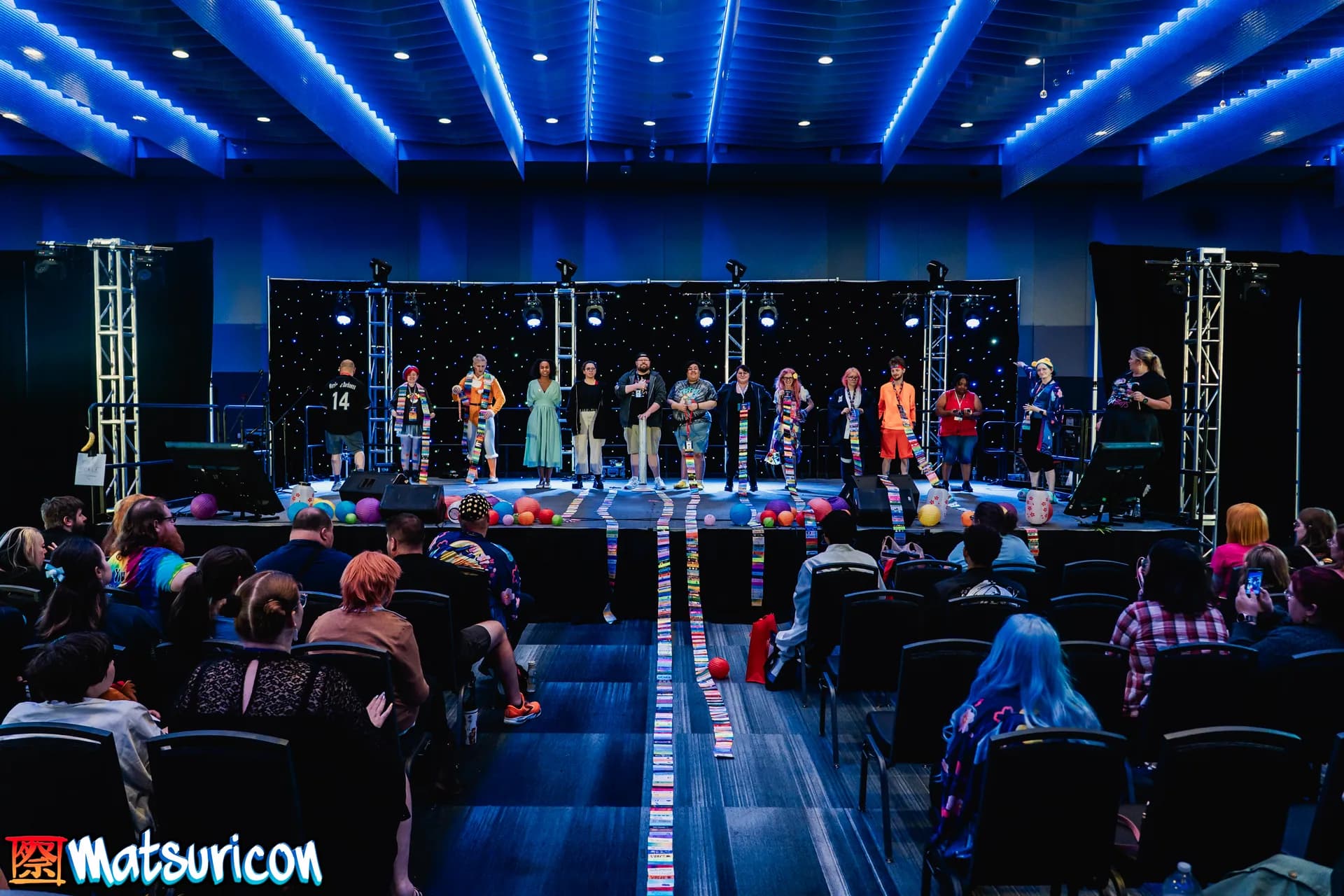 Ribbon collectors stand across a Matsuricon stage, many wearing floor-length chains of colorful badge ribbons, while an audience watches.